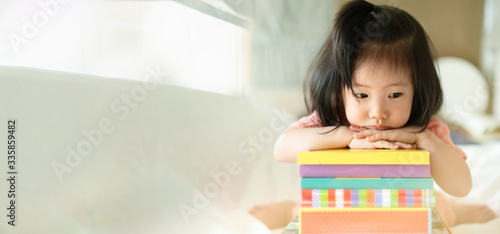 Panoramic of adorable toddler girl looking downward and feeling bored and unhappy while sitting with the book stack.