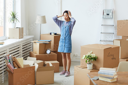 Full length portrait of frustrated Asian woman panicking while standing among cardboard boxes in empty room, house moving or relocation concept, copy space
