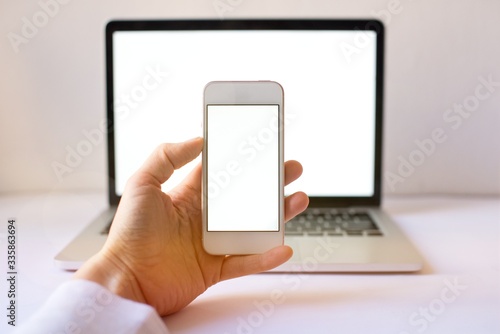 Close-up of smartphone with blank screen in hands of young woman sitting at white table and touching screen. On table laptop with blank screen,cup of coffee. White style. Minimal. Mock up,space for ad