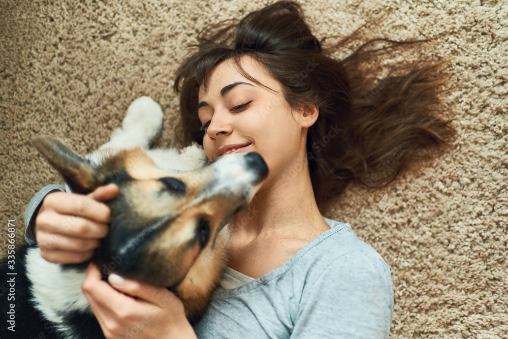 Portrait young joyful woman lying on carpet with cute pet. Happy girl playing and stroking Welsh Corgi dog. Concept stay at home, friendship with dog, happy moments. Portrait young joyful woman lying on carpet with cute pet. Happy girl playing and stroking Welsh Corgi dog. Concept stay at home, friendship with dog, happy moments.