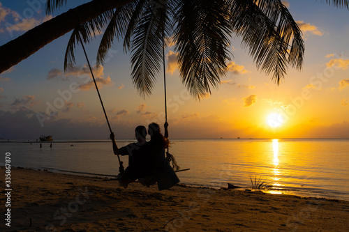Silhouetted couple in love walks on the beach during sunset. Riding on a swing tied to a palm tree and watching the sun go down into the ocean