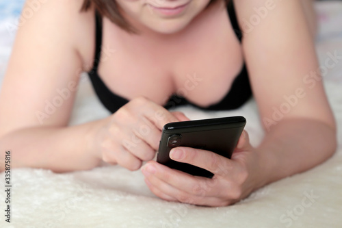 Woman in black bra lying on a bed and using smartphone. Mobile phone in female hands, concept of online addiction, sms, social media, adultery