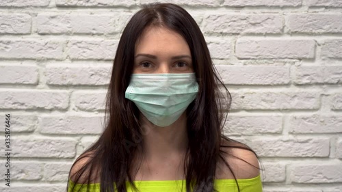 Young beautiful Caucasian brunette woman with long hair and brown eyes in turquoise protective medical mask from virus, laughs and smiles on white brick wall background close up. Girl is happy. photo