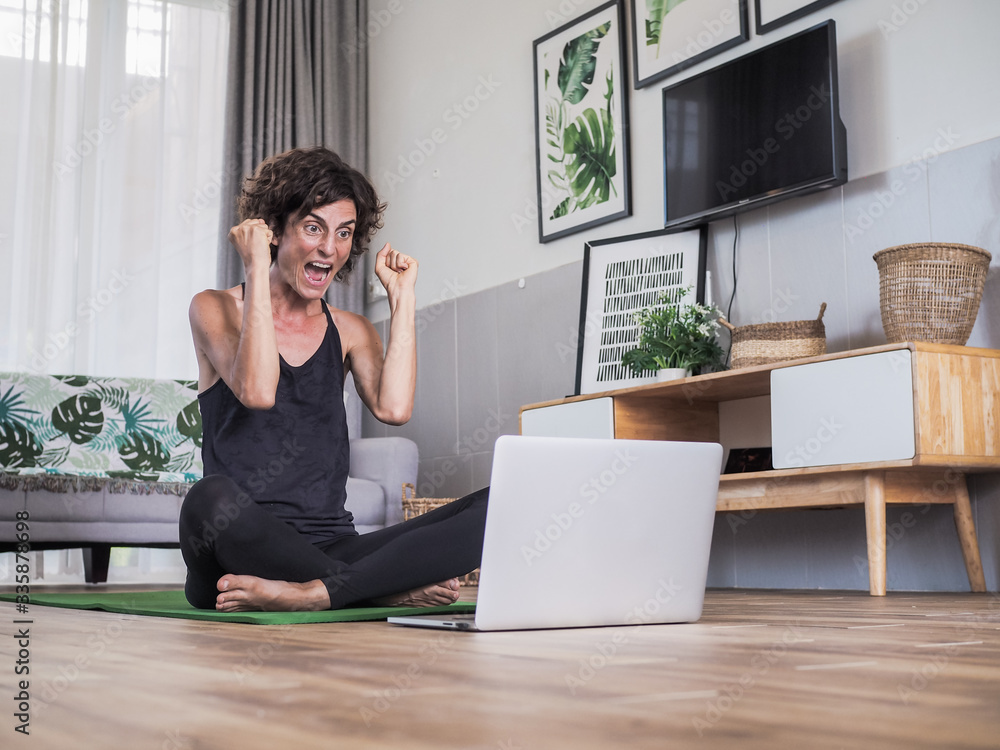 happy excited and smiling women in winner pose sitting crossed legged on the floor on a yoga mat in her living room at home looking at laptop having a video conference happy excited and smiling women in winner pose sitting crossed legged on the floor on a yoga mat in her living room at home looking at laptop having a video conference