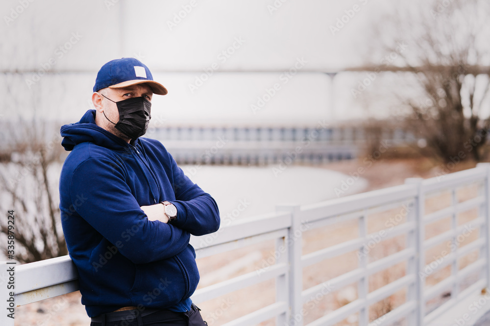 Man in a dark blue hoodie, dark blue baseball cap with a mask on his face is leaning against a fence looking with concern, he is worried about a virus problem. Man in a dark blue hoodie, dark blue baseball cap with a mask on his face is leaning against a fence looking with concern, he is worried about a virus problem.