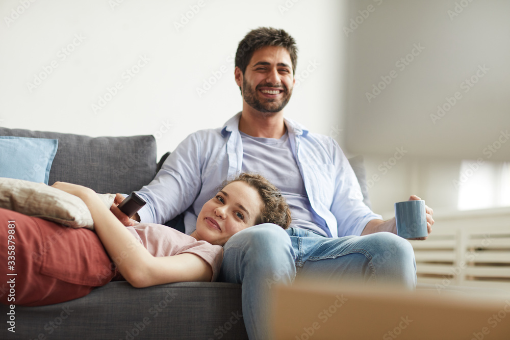 Portrait of happy couple watching TV together while sitting on sofa at home in cozy apartment enjoying lazy time, copy space
