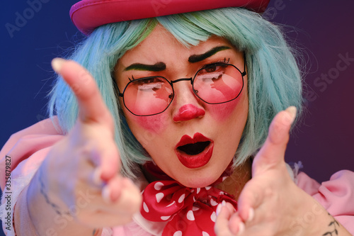 Close-up portrait of a clown girl with blue hair in a pink hat, pink dress and glasses. A clown shows different human emotions. 