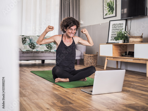 happy excited and smiling women in winner pose sitting crossed legged on the floor on a yoga mat in her living room at home looking at laptop having a video conference 