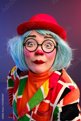 Close-up portrait of a clown girl with make-up, blue hair, a red hat, a colored checkered jacket and glasses. 