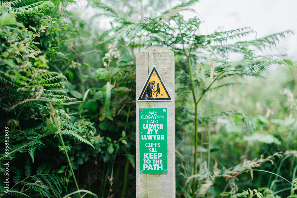 Cliffs kill, keep to the path tourist sign in welsh and english languages on the coastal pathway in Wales, UK. Cliffs kill, keep to the path tourist sign in welsh and english languages on the coastal pathway in Wales, UK.