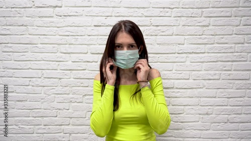 Cute Caucasian brunette woman with long hair in green jacket put on a turquoise protective medical mask from virus on white brick wall background close up. Covid 19 coronavirus protection concept. photo