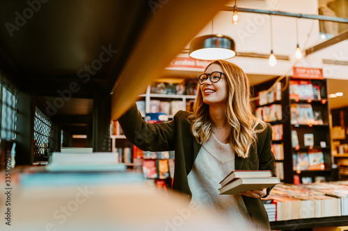 Photo of young blonde woman with glasses looking at book shelves at bookstore. Young woman is looking for new books to buy. photo