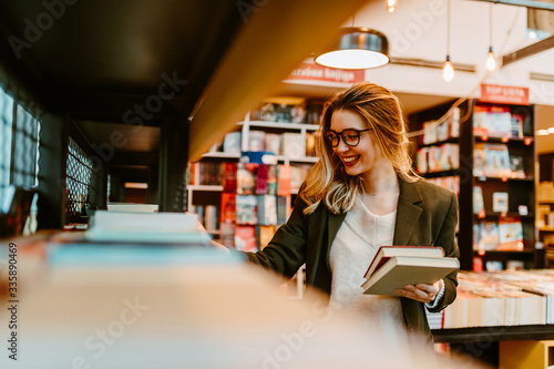 Portrait of young blonde woman with beautiful smile trying to find new books to read. Young woman is looking at book shelves in order to find something interesting for her to read. photo
