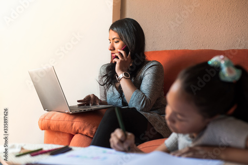 Latin young woman working at home calling with the mobile phone using the laptop while her daughter are doing the homework. Telecommuting concept during covid infection.