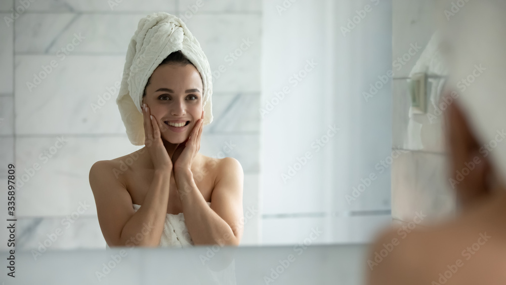Smiling young woman in bath towel look in mirror after shower satisfied with healthy glowing facial skin, happy millennial girl touch clean face after beauty procedures or treatment, skincare concept Smiling young woman in bath towel look in mirror after shower satisfied with healthy glowing facial skin, happy millennial girl touch clean face after beauty procedures or treatment, skincare concept