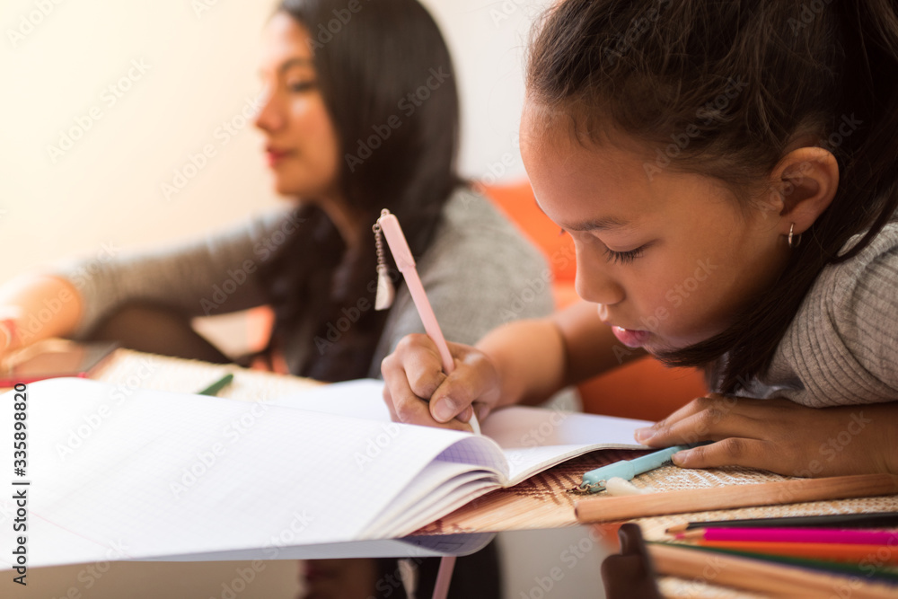 Little latin girl writing on a notebook at home while her mother watches over her. Concept of telecommuting and homework with tablet and online technologies. Little latin girl writing on a notebook at home while her mother watches over her. Concept of telecommuting and homework with tablet and online technologies.