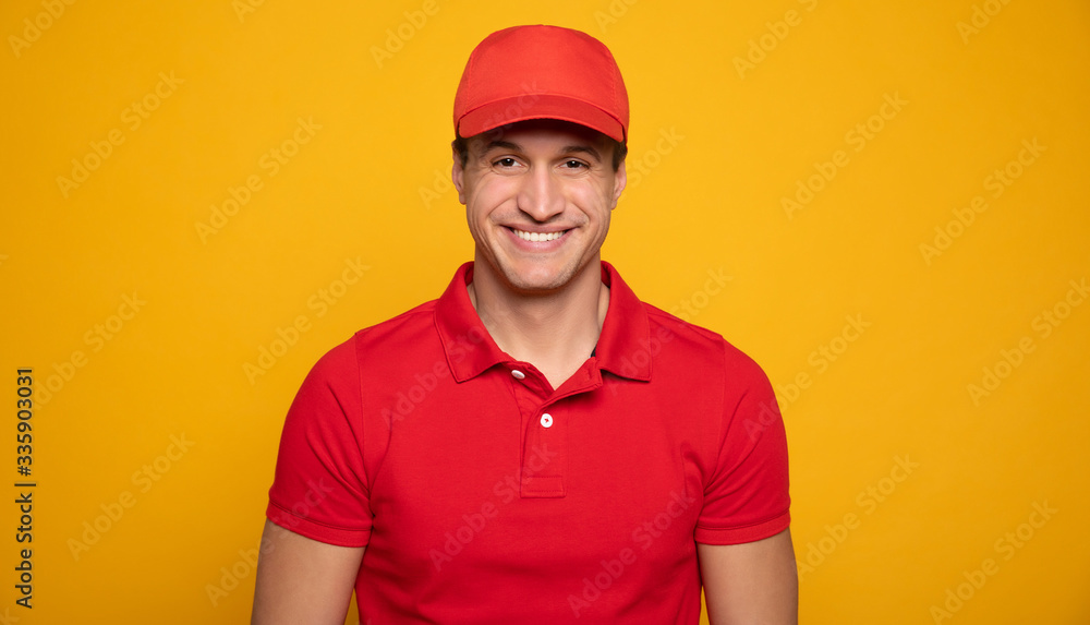 Handsome young happy courier or delivery man in red uniform is posing isolated on yellow background.