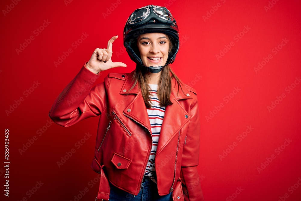 Young beautiful brunette motocyclist woman wearing motorcycle helmet and red jacket smiling and confident gesturing with hand doing small size sign with fingers looking and the camera. Measure Young beautiful brunette motocyclist woman wearing motorcycle helmet and red jacket smiling and confident gesturing with hand doing small size sign with fingers looking and the camera. Measure