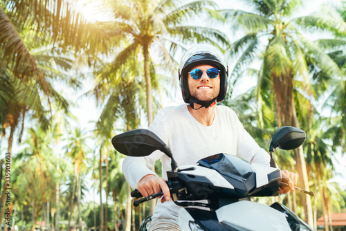 Happy smiling male tourist in helmet and sunglasses riding motorbike scooter during his tropical vacation under palm trees.