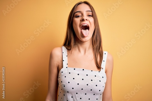Valokuva Young beautiful brunette woman wearing casual dress standing over yellow background sticking tongue out happy with funny expression
