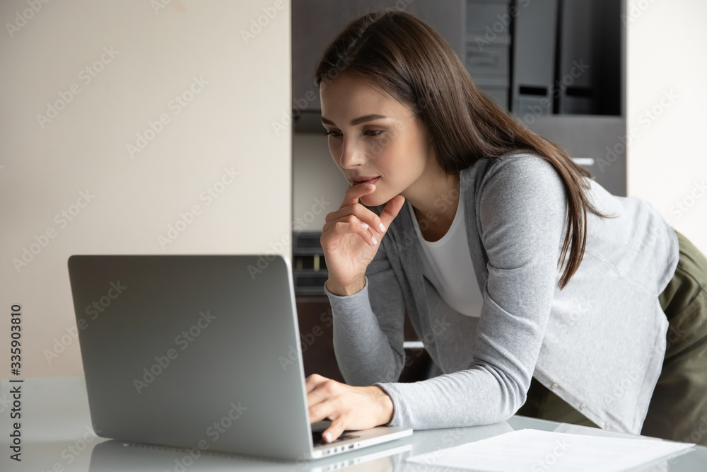Pensive millennial female employee stand at desk in modern office look at laptop screen browsing Internet wireless connection, focused young woman busy working on computer, consult client online Pensive millennial female employee stand at desk in modern office look at laptop screen browsing Internet wireless connection, focused young woman busy working on computer, consult client online
