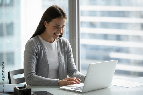 Smiling millennial girl sit at desk look at laptop screen working in office, happy young woman employee typing texting consulting business client or customer online on modern computer gadget