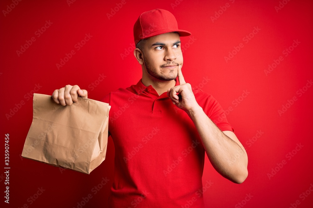 Young handsome delivery man holding paper bag with takeaway food over red background serious face thinking about question, very confused idea Young handsome delivery man holding paper bag with takeaway food over red background serious face thinking about question, very confused idea
