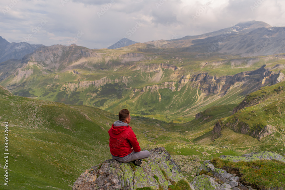 Young man in a red jacket on the stones looks at a magnificent view of the Alps mountains. Young man in a red jacket on the stones looks at a magnificent view of the Alps mountains.