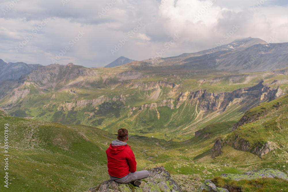 Young man in a red jacket on the stones looks at a magnificent view of the Alps mountains. Young man in a red jacket on the stones looks at a magnificent view of the Alps mountains.