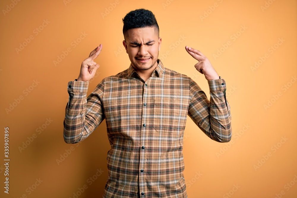 Young handsome man wearing casual shirt standing over isolated yellow background gesturing finger crossed smiling with hope and eyes closed. Luck and superstitious concept. Young handsome man wearing casual shirt standing over isolated yellow background gesturing finger crossed smiling with hope and eyes closed. Luck and superstitious concept.