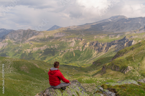 Young man in a red jacket on the stones looks at a magnificent view of the Alps mountains. © fotoplaton