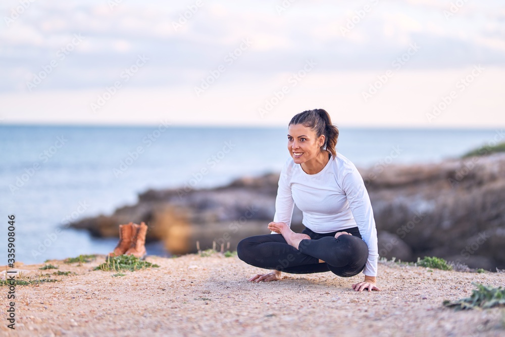 Young beautiful sportwoman smiling happy practicing yoga. Coach with smile on face teaching crow pose at the beach