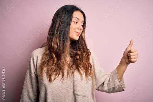 Young beautiful brunette woman wearing casual sweater standing over pink background Looking proud, smiling doing thumbs up gesture to the side