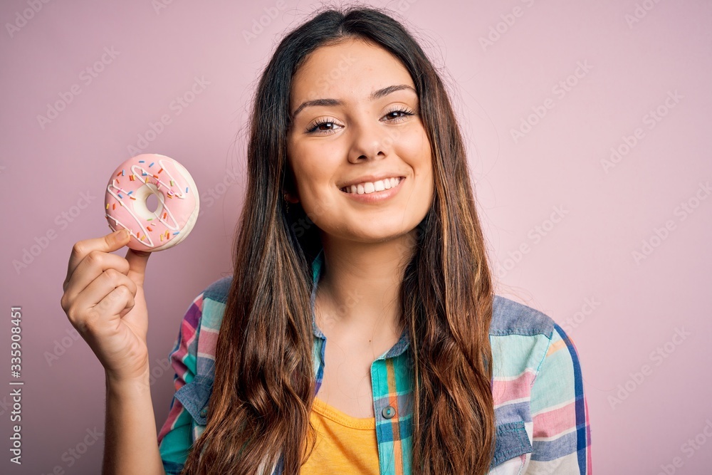 Young beautiful brunette woman eating sweet pink doughnut over isolated background with a happy face standing and smiling with a confident smile showing teeth Young beautiful brunette woman eating sweet pink doughnut over isolated background with a happy face standing and smiling with a confident smile showing teeth