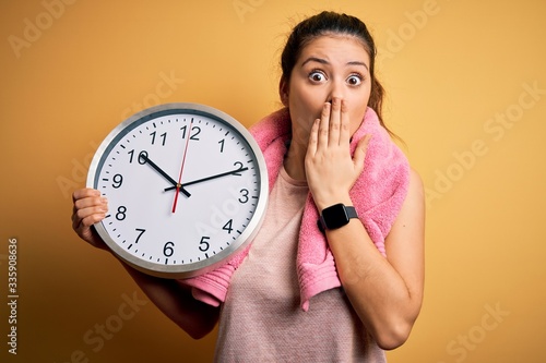 Young beautiful brunette sportswoman wearing towel doing countdown using big clock cover mouth with hand shocked with shame for mistake, expression of fear, scared in silence, secret concept