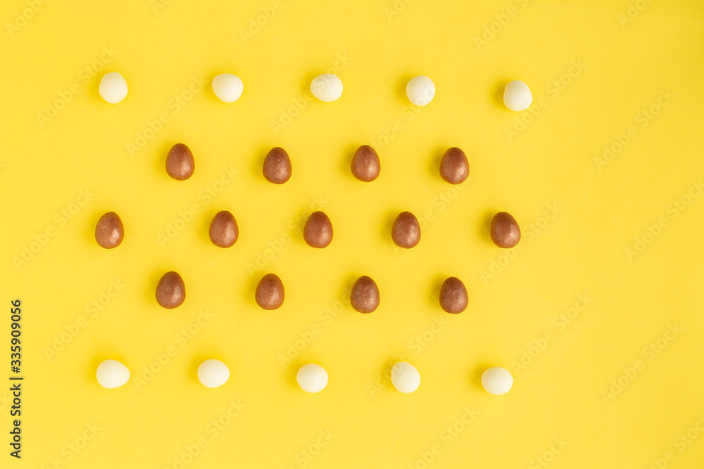 Several rows of small eggs made of white and dark chocolate on yellow background.Easter greeting card, symbol of celebration Sunday of Christ.Holiday composition, copy space for text congratulations