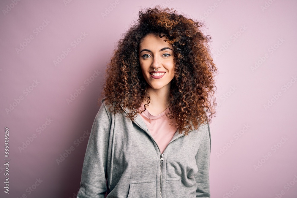 Young beautiful brunette sportswoman with curly hair and piercing wearing sportswear with a happy and cool smile on face. Lucky person.