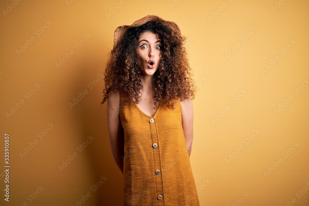 Beautiful brunette woman on vacation with curly hair and piercing wearing hat and dress afraid and shocked with surprise expression, fear and excited face. Beautiful brunette woman on vacation with curly hair and piercing wearing hat and dress afraid and shocked with surprise expression, fear and excited face.