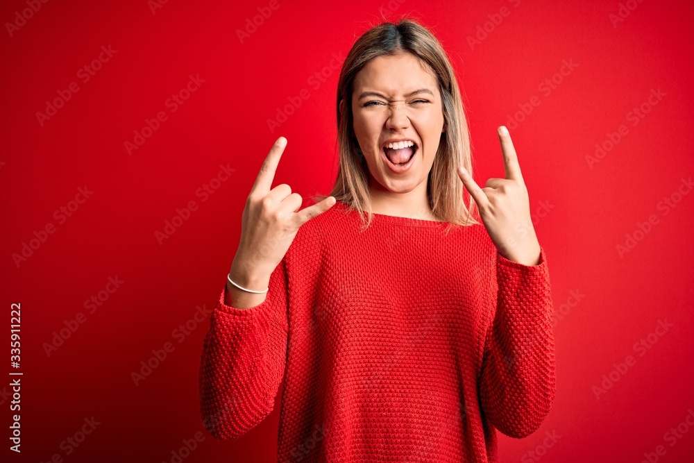 Young beautiful blonde woman wearing casual sweater over red isolated background shouting with crazy expression doing rock symbol with hands up. Music star. Heavy music concept. Young beautiful blonde woman wearing casual sweater over red isolated background shouting with crazy expression doing rock symbol with hands up. Music star. Heavy music concept.