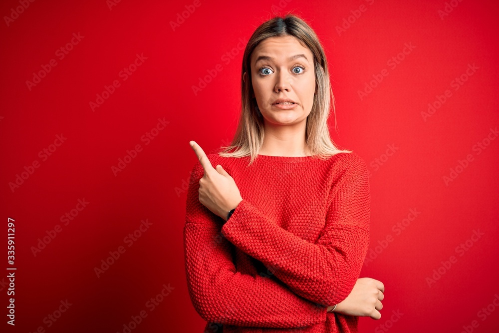 Young beautiful blonde woman wearing casual sweater over red isolated background Pointing aside worried and nervous with forefinger, concerned and surprised expression Young beautiful blonde woman wearing casual sweater over red isolated background Pointing aside worried and nervous with forefinger, concerned and surprised expression