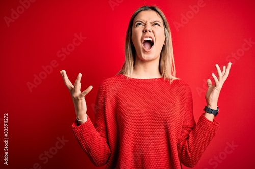 Young beautiful blonde woman wearing casual sweater over red isolated background crazy and mad shouting and yelling with aggressive expression and arms raised. Frustration concept.