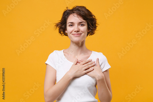 Smiling young brunette woman girl in white t-shirt posing isolated on yellow orange wall background studio portrait. People lifestyle concept. Mock up copy space. Holding hands folded on chest, heart. photo