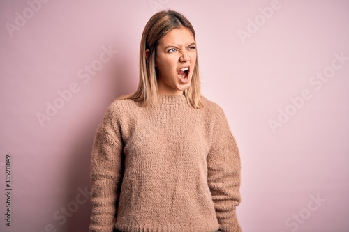 Young beautiful blonde woman wearing winter wool sweater over pink isolated background angry and mad screaming frustrated and furious, shouting with anger. Rage and aggressive concept.