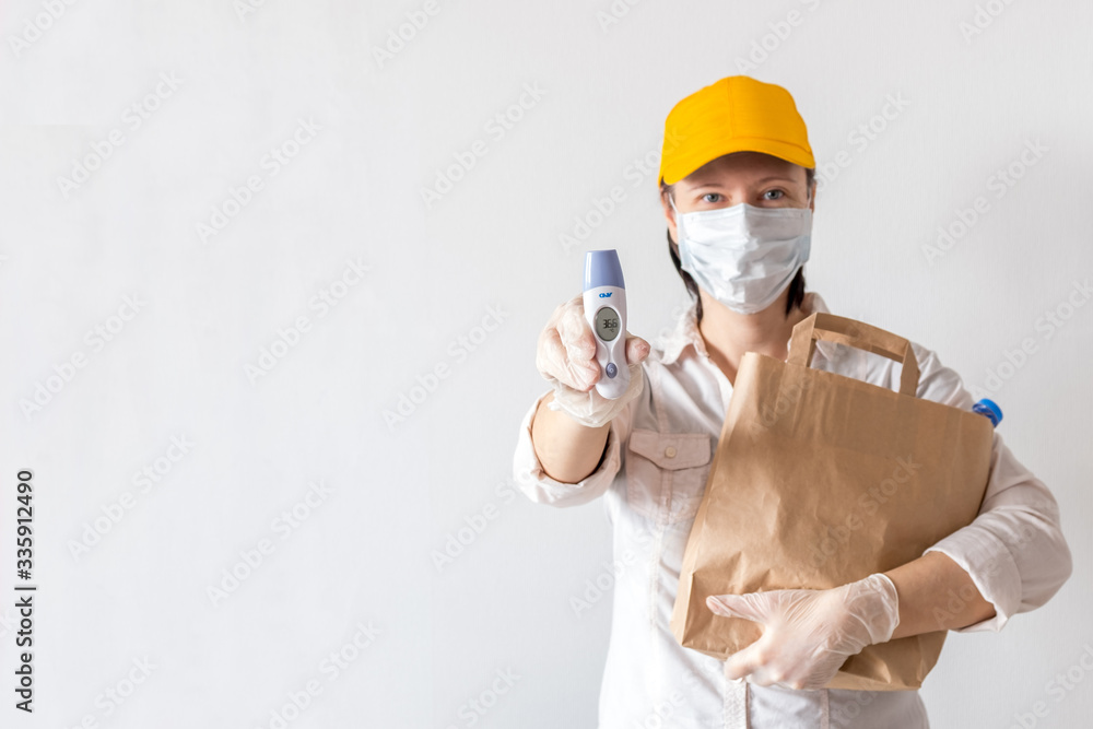 Delivery woman holding a paper bag with food on a white background. food delivery woman in a protective mask and gloves. Delivery holds an electronic thermometer and shows that it is healthy. Delivery woman holding a paper bag with food on a white background. food delivery woman in a protective mask and gloves. Delivery holds an electronic thermometer and shows that it is healthy.