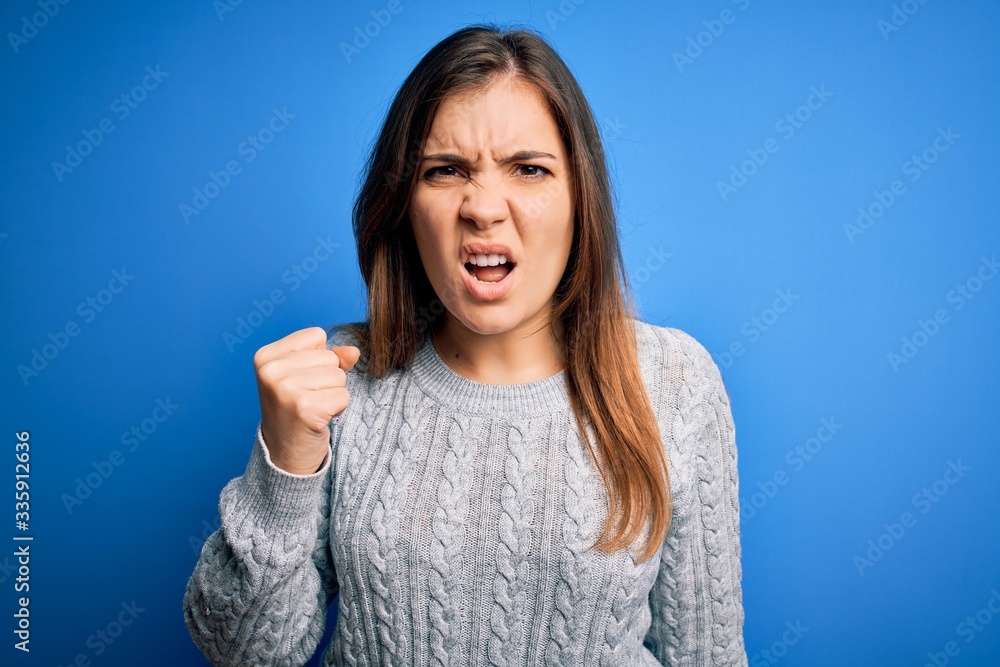 Beautiful young woman wearing casual wool sweater standing over blue isolated background angry and mad raising fist frustrated and furious while shouting with anger. Rage and aggressive concept. Beautiful young woman wearing casual wool sweater standing over blue isolated background angry and mad raising fist frustrated and furious while shouting with anger. Rage and aggressive concept.
