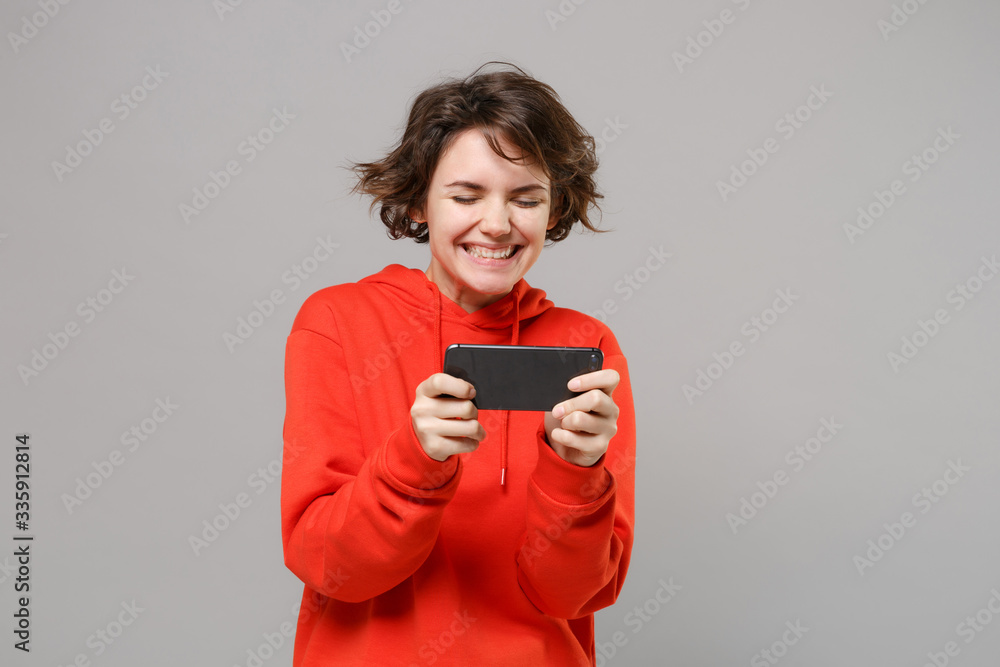 Cheerful young brunette woman girl in casual red hoodie posing isolated on grey background studio portrait. People sincere emotions lifestyle concept. Mock up copy space. Play game with mobile phone. Cheerful young brunette woman girl in casual red hoodie posing isolated on grey background studio portrait. People sincere emotions lifestyle concept. Mock up copy space. Play game with mobile phone.