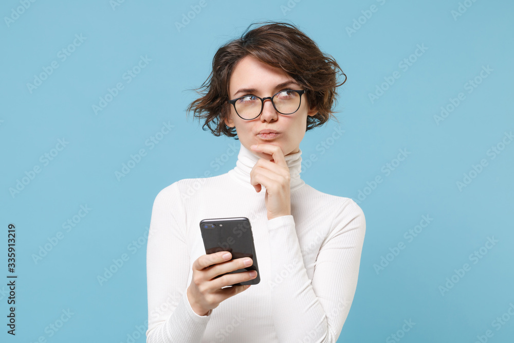 Pensive young brunette woman girl in casual white clothes, eyeglasses isolated on pastel blue background. People lifestyle concept. Mock up copy space. Using mobile phone, put hand prop up on chin. Pensive young brunette woman girl in casual white clothes, eyeglasses isolated on pastel blue background. People lifestyle concept. Mock up copy space. Using mobile phone, put hand prop up on chin.