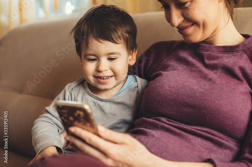 close up portrait of a pregnant caucasian mother spending time with her little boy using a phone on the sofa photo