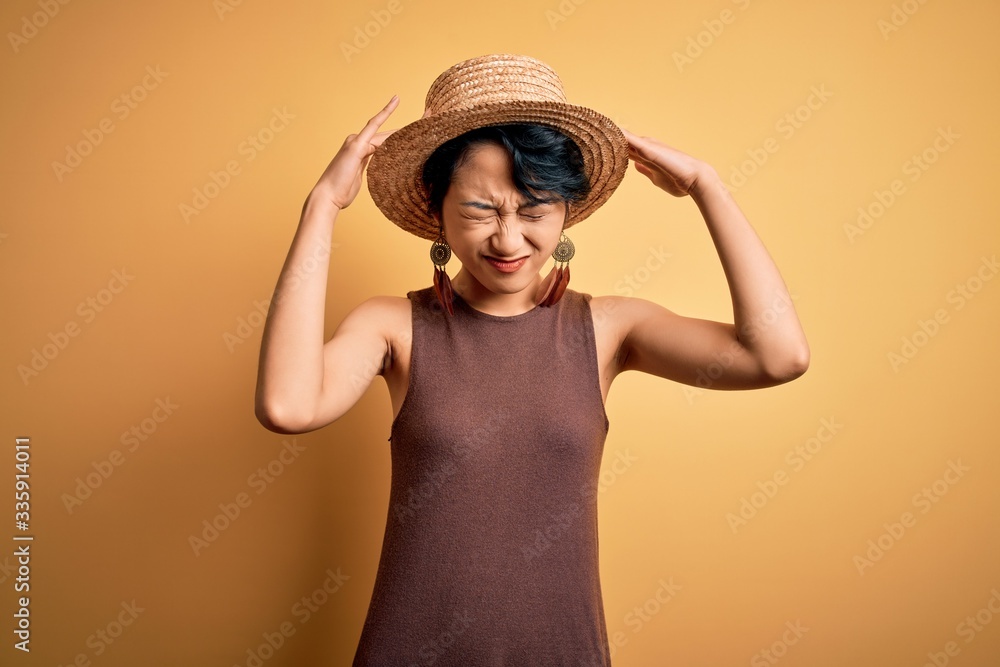 Young beautiful asian girl wearing casual t-shirt and hat over isolated yellow background suffering from headache desperate and stressed because pain and migraine. Hands on head. Young beautiful asian girl wearing casual t-shirt and hat over isolated yellow background suffering from headache desperate and stressed because pain and migraine. Hands on head.