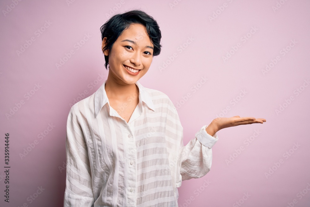 Young beautiful asian girl wearing casual shirt standing over isolated pink background smiling cheerful presenting and pointing with palm of hand looking at the camera. Young beautiful asian girl wearing casual shirt standing over isolated pink background smiling cheerful presenting and pointing with palm of hand looking at the camera.